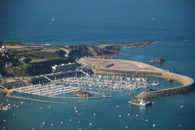 a rocky headland sheltering a curved breakwater inside of which is a large marina with many masted boats inside