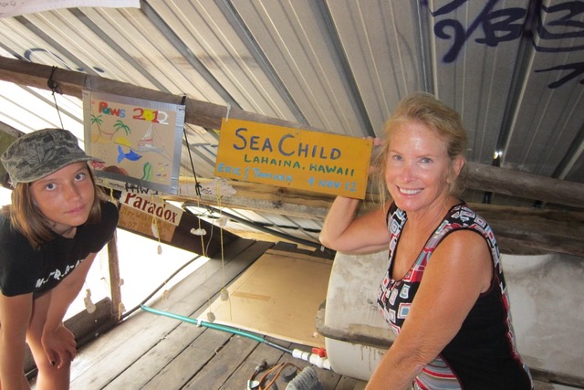 A woman on the right in front of a plaque with the boats name in it and a girl opposite inside a makeshift shelter on the beach