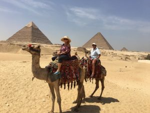 two camels with people riding them in front of a line of pyramids