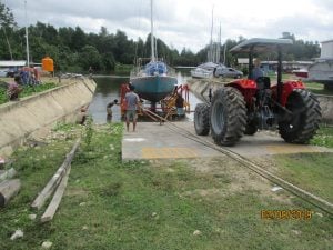 red tractor in the foreground parked over railway rails and a blue boat on a trailer being pulled up the ramp from the river