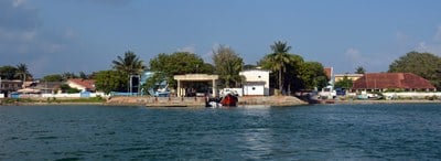 a shoreline with a few trees and a palm tree, brown roofed buildings and a concrete pier with a concrete arched structure on top of it and a white house