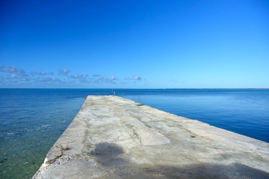 a low concrete dock extending out into a blue sea with small sticks marking a channel for approach