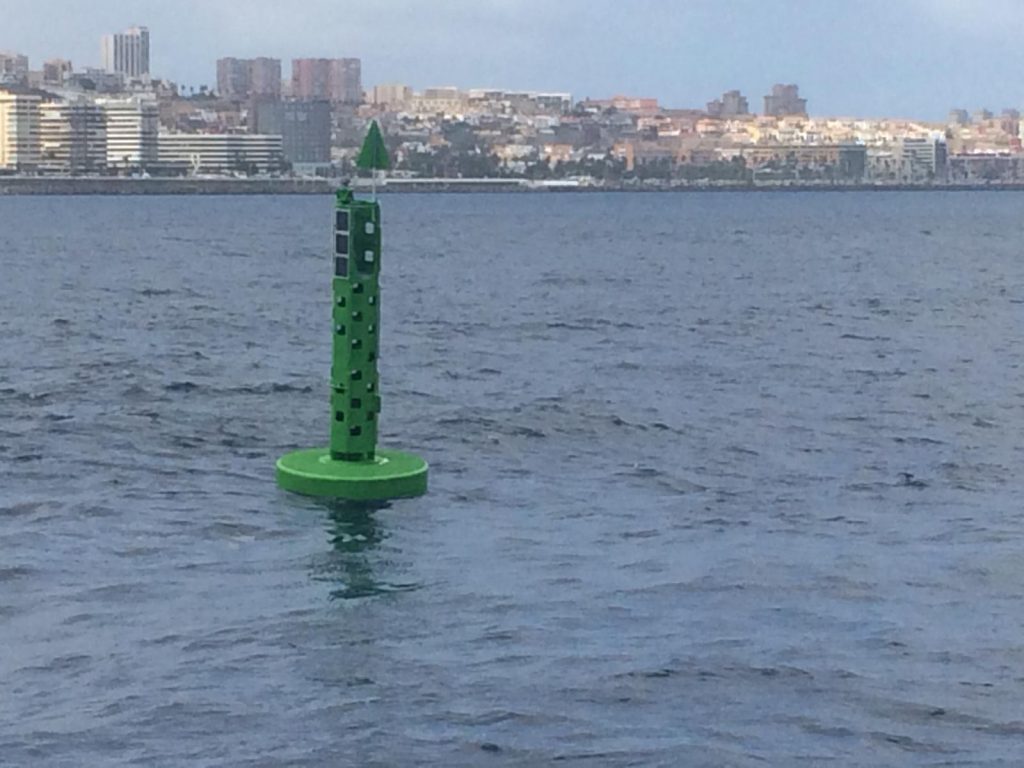 green buoy shaped like a narrow cylinder with a green upward pointing triangle on top in the sea with las palmas city behind