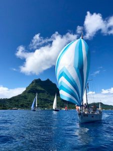yacht with spinnaker flying sailing towards the camera with the rest of the fleet behind as they leave the island of bora bora in french polynesia