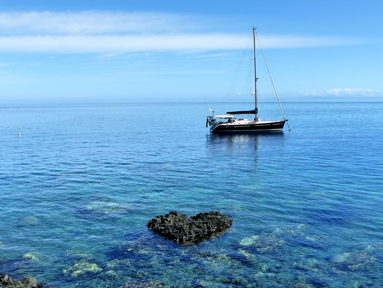 yacht at anchor in crystal clear water with a reef in the foreground and a rock shaped like a heart