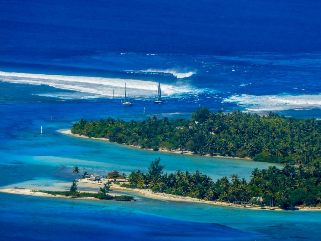 aerial photo of the reef pass with breaking waves, a small motu and 2 yachts entering in to the calm blue waters of the lagoon