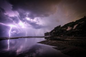 purple filter showing coastal cliffs and shallow water running up to beach with dramatic lightning and clouds over the sea in the distance