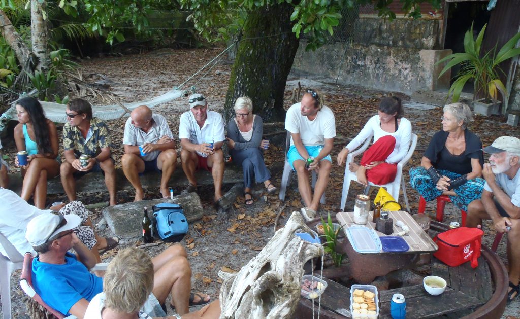 a circle of people sitting on logs and plastic chairs, chatting and laughing some with drinks in their hands and others not
