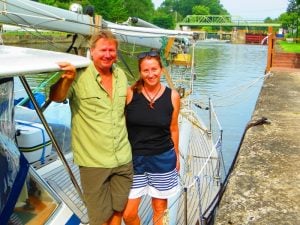 sailing couple both in shorts with a tan standing on the side of their boat smiling with a canal extending behind.