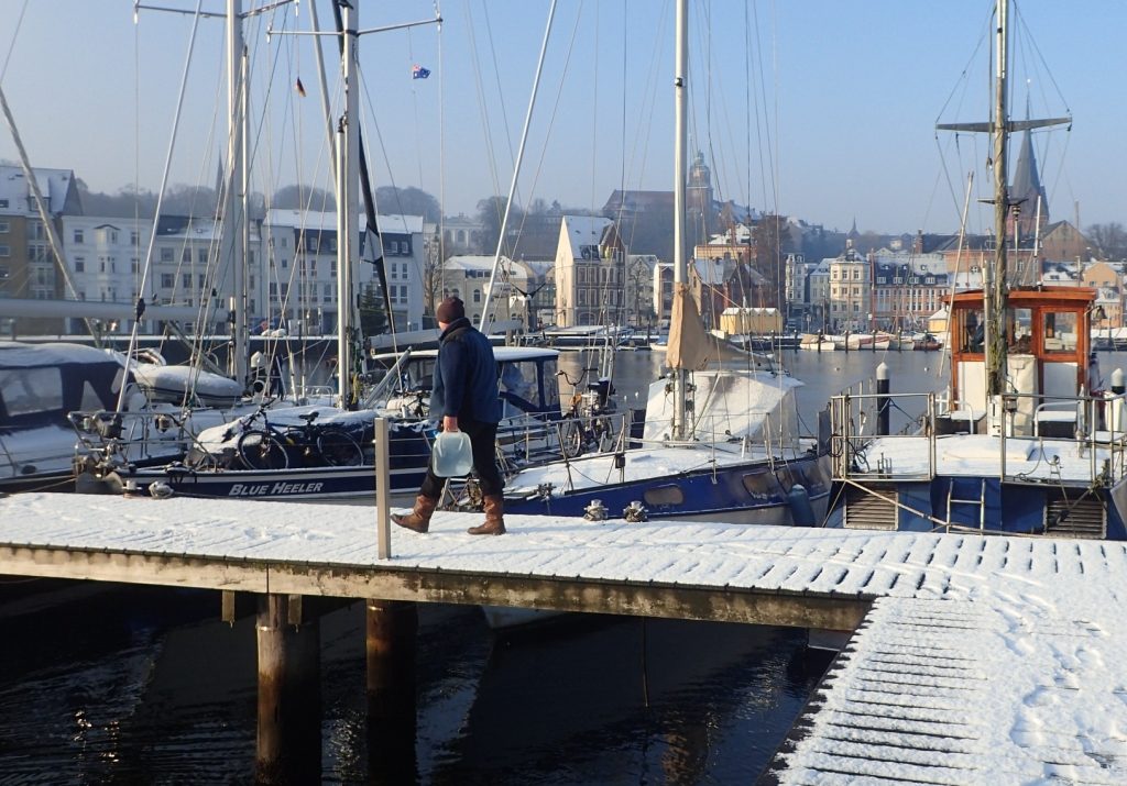 a man dressed in warm clothes, boots and a wooly hat carries water jerry cans along a snow laden wooden pontoon with boats tied to it, also covered in snow