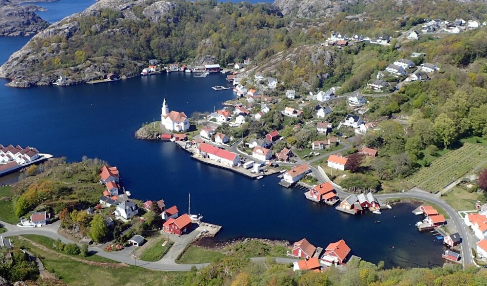 aerial view of a bright blue fjord with a conspicuous village and church spire showing one lone yacht in the harbour