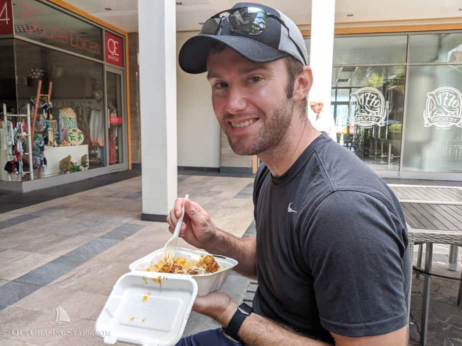 a man with a cap and sunglasses pushed up smiling and eating local takeaway food out of a polystirine dish with a plastic fork