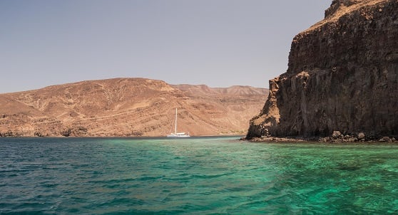 high rock walls and turquoise sea with an arid landscape and a white yacht at anchor in the middle