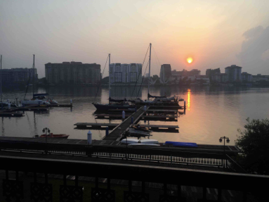 a view at dusk over the river with the marina in the foreground and the city in the background on the opposite shore with a red sun setting behind