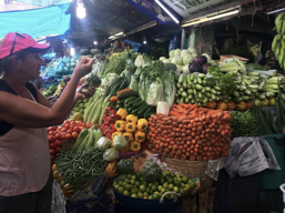 A lady in a cap passing money over a market stall full of vegetables of all colours to the lady store holder