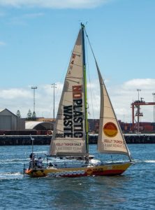 yellow yacht covered in logos with a white mainsail and the words no plastic waste running down it and a white front sail with the no plastic waste logo and Jon at the helm