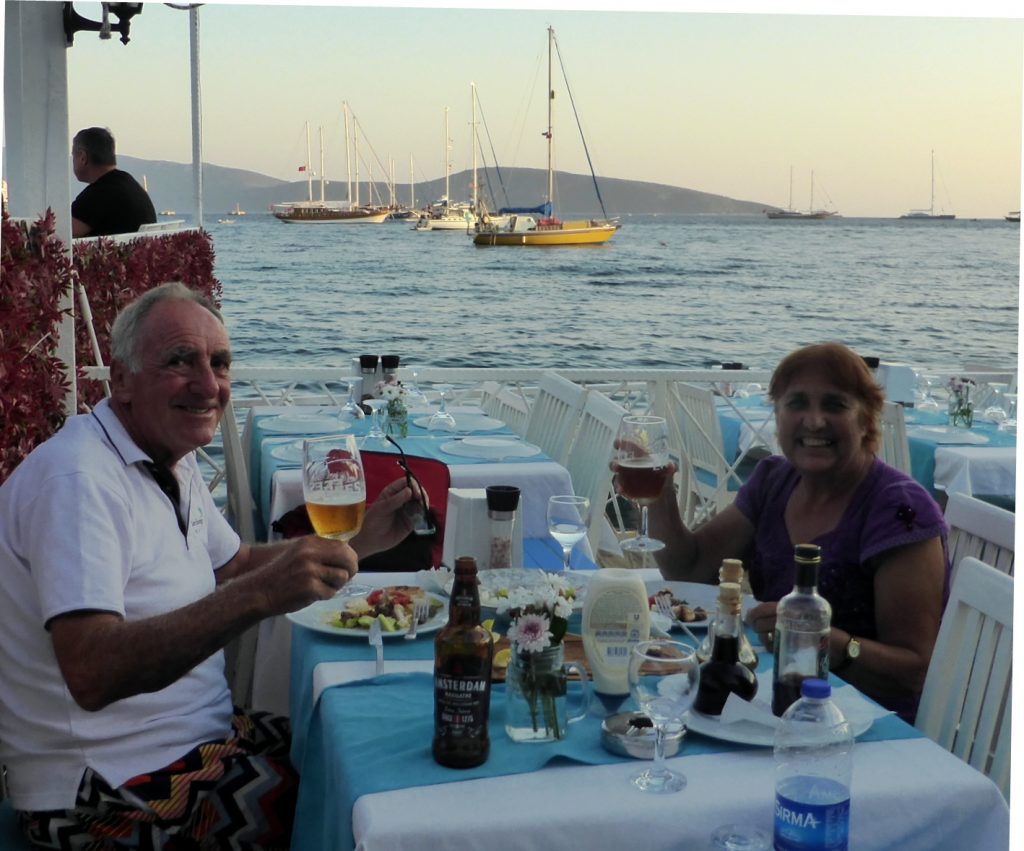 man and woman sitting at a waterside outdoor restaurant raising a glass with their boat with a yellow hull in the background