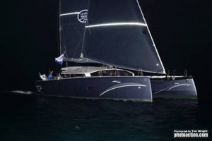 night time with a dark sky and dark seas and a beautiful silver catamaran with its twin hulls close to the camera on port tack sailing close hauled across the line