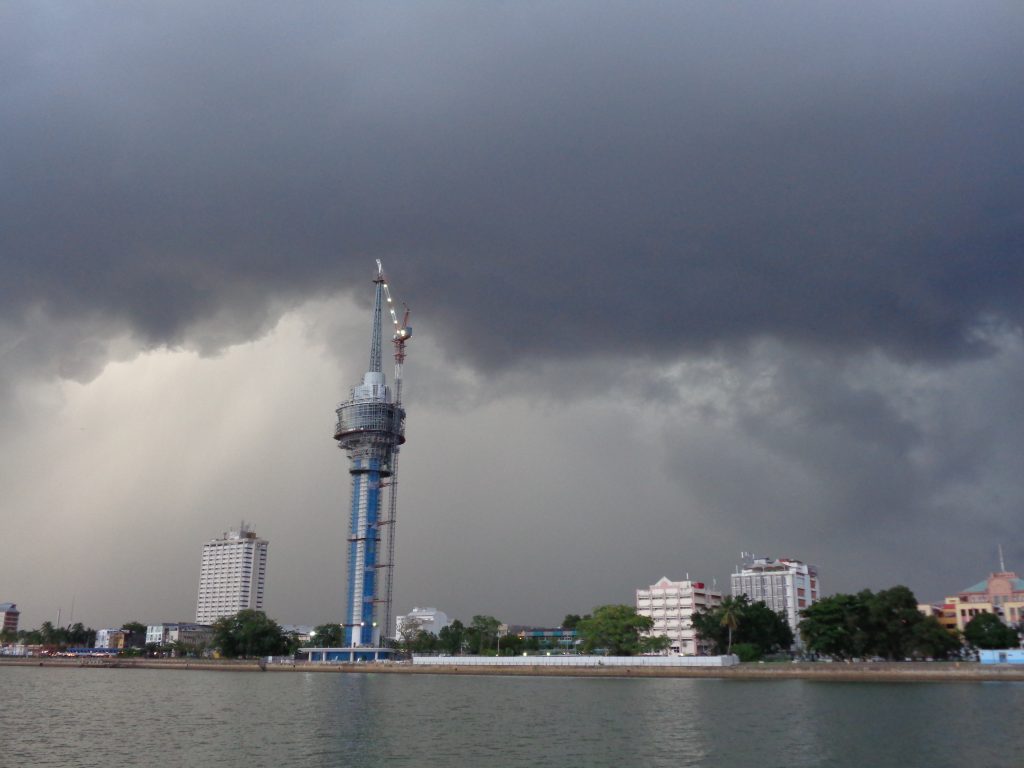 a skyline with a tall water tower and three blocks of flats, with grey flat water in front and dramatic grey storm clouds behind