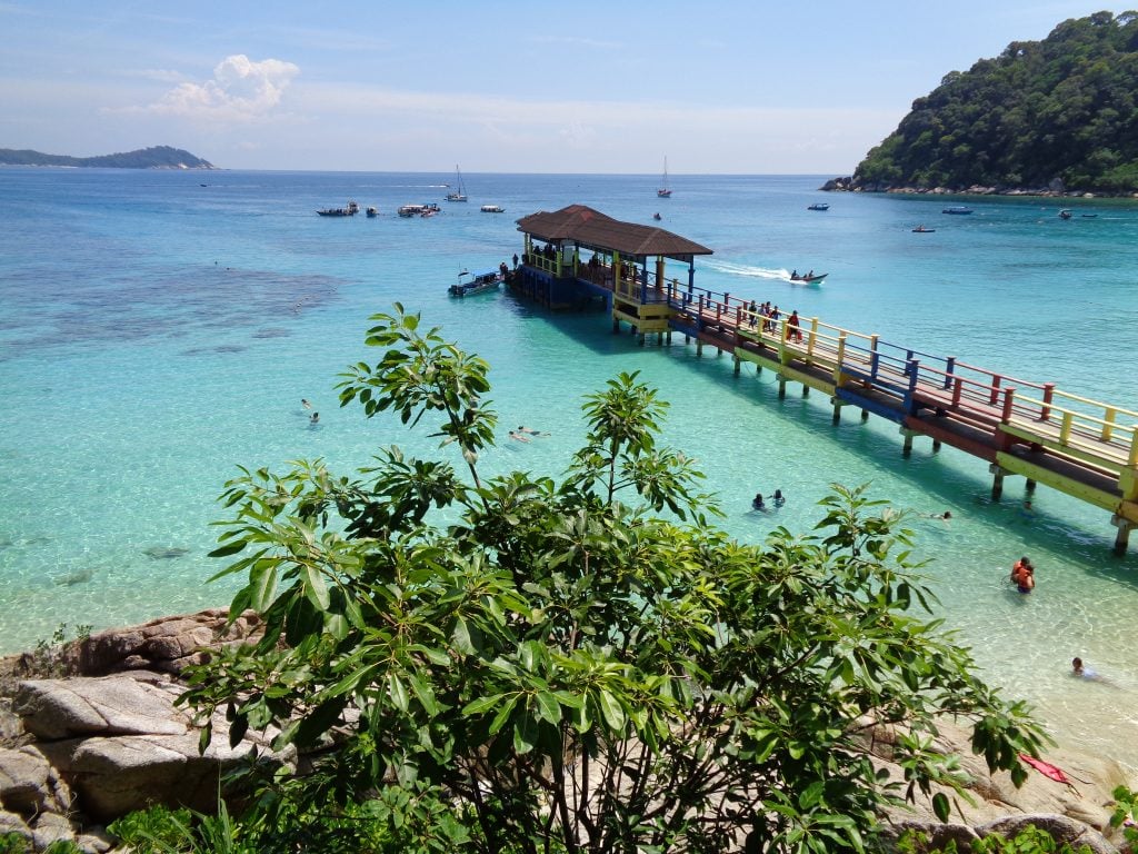turquoise waters with a view from a beach and a tree in the foreground, then a long wooden jetty with a covered portion at the end extending out into the sea