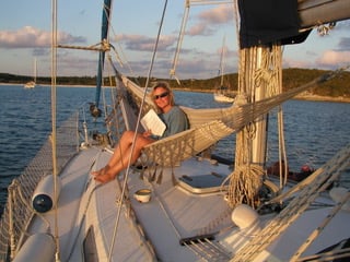 a lady sitting sideways on a hammock hung on the foredeck of a boat holding some papers