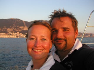 a woman and man smiling at the camera wearing white shirts and blue jumpers with land and sea behind them