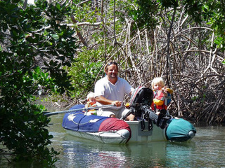 an inflatable dinghy with its outboard raised in a narrow mangrove river with dad in the middle rowing and 2 children with blonde hair accompanying him