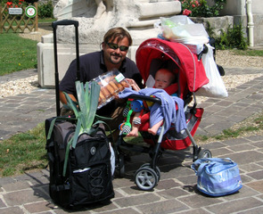 a man with his child in a pushchair, he crouching down and in front of them a wheelie shopping trolley like a suitcase packed with groceries