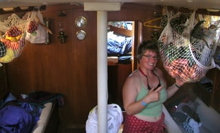 a smiling lady in sleeveless top and shorts with sunglasses hanging around her neck down below in the saloon of the boat surrounded by white nets hanging from the roof containing fruits and vegetables