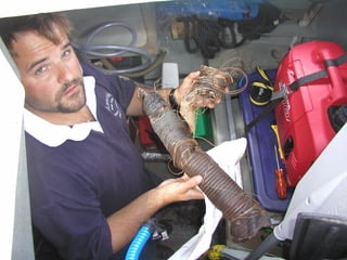 a man with a beard in an engine room holding a length of broken hose and a spring in his hands