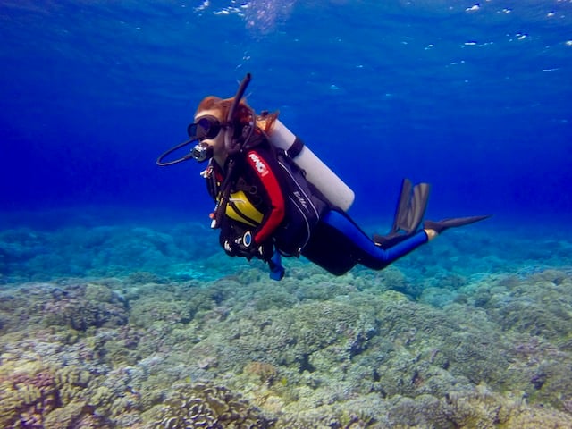 a diver with long red hair and fully equipped swimming over a reef in blue sea