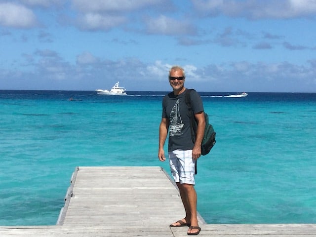 a man in a dark t shirt and white shorts with a backpack standing at the root of a wooden dock with blue blue sea beyond and his white motorboat anchored some distance away in darker blue water