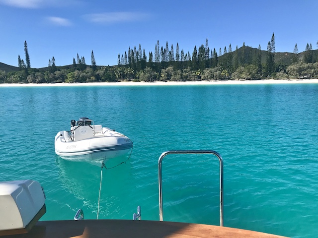 view from the back of the boat with the white dinghy floating off the back on top of crystal clear water and a strip of white sand ashore topped with tall narrow pine trees in a long line like soldiers