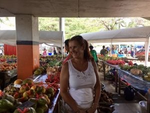 a lady with red hair wearing a white sleeveless top in the middle of an outdoor market full of fruits and vegetables