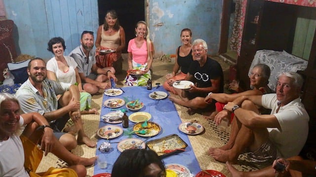 A group of approximately 10 people sitting on the floor dressed in hot weather clothes sitting around a blue rectangular cloth laden with dishes of food