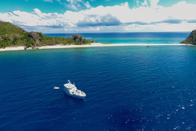 blue water, a green island with a white sand spit in the distance and a white motor yacht anchored