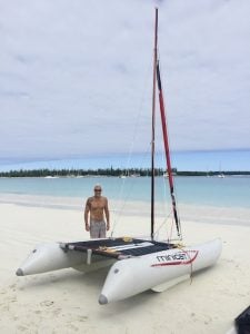 a man standing on a white sand beach with an inflatable white dinghy catamaran with one mast and a red foresail