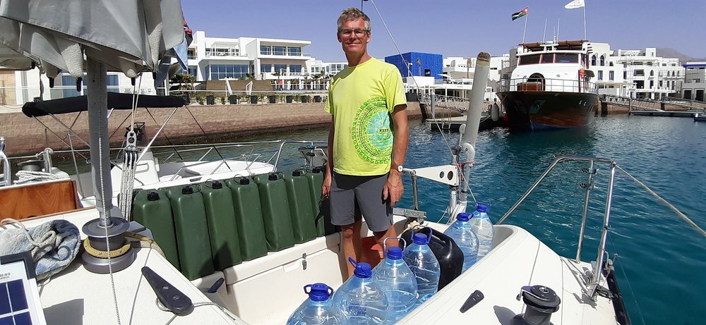 a man with grey hair and glasses and a tan wearing a yellow t shirt and grey shorts standing in teh cockpit of his boat with a large number of green jerry cans containing fuel on his right cockpit seats and many large water bottles on his left cockpit seat