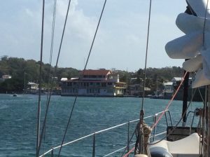 view from the deck of a boat across an anchorage to a town on shore with a tall house with a red roof and other smaller buildings next to it