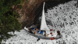 looking down on a yacht with a small sail up trapped up against the cliffs with lots of white spume from the waves and items in the water around it