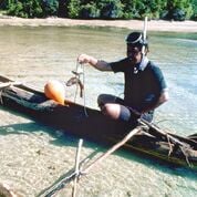 a fisherman wearing a snorkel and mask in a black shirt is holding up a fish on a line as he sits in a narrow canoe with outriggers