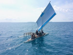 a traditional sailing canoe with several men on board and a square white sail