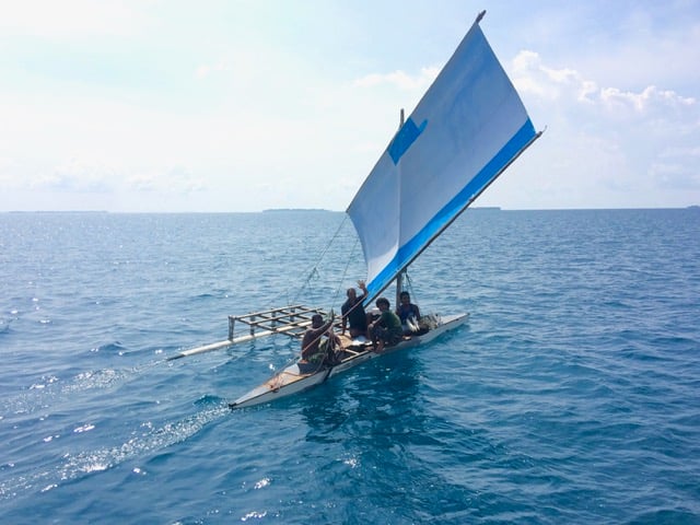 a traditional sailing canoe with several men on board and a square white sail