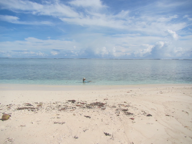a white almost pink sandy beach in the foreground leading down to the shallows and flat calm sea stretching all the way to the horizon with a blue sky with wispy clouds