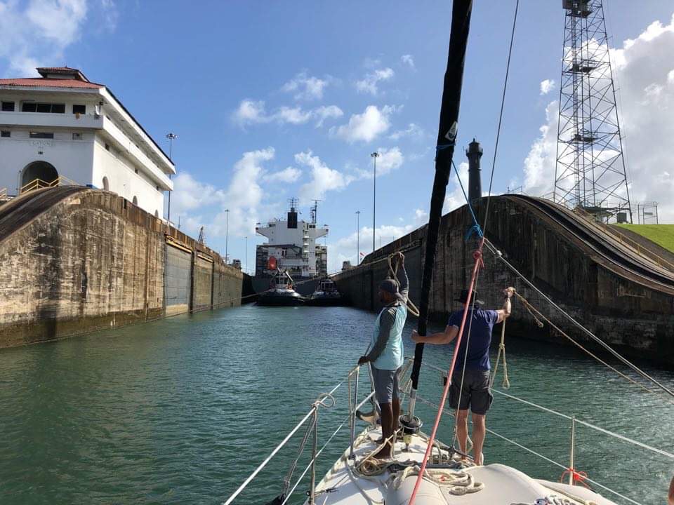 a boat entering a lock with a large ship ahead and the high canal walls either side with 2 men on the foredeck holding lines