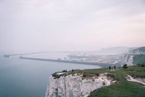 tall white cliffs with grass on top and people looking out to sea with a view of Dover harbour with large breakwaters forming a protected large harbor