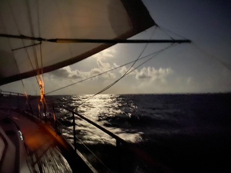 a view up the starboard side of a yacht with a poledout genoa and the sun behind the sail casting a shine on the sea