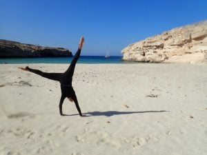 a wetsuit clad woman cartwheeling on a white sand beach with blue sea in the background