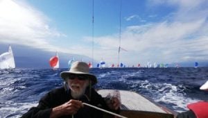 Man at the helm of a boat with yachts behind him with spinnakers flying.